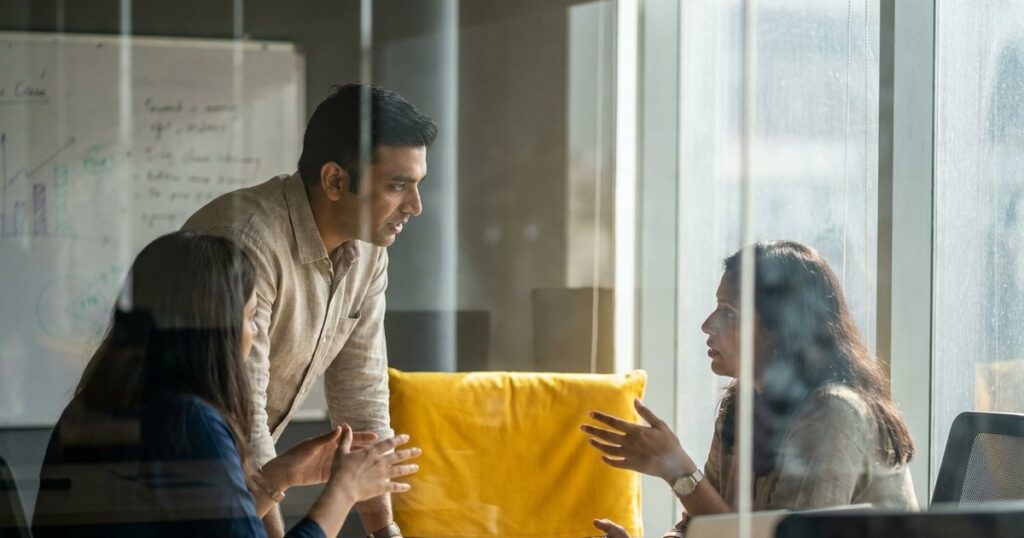Indian founders discussing AI tools for negotiation in a sunlit office, seen through a glass partition.