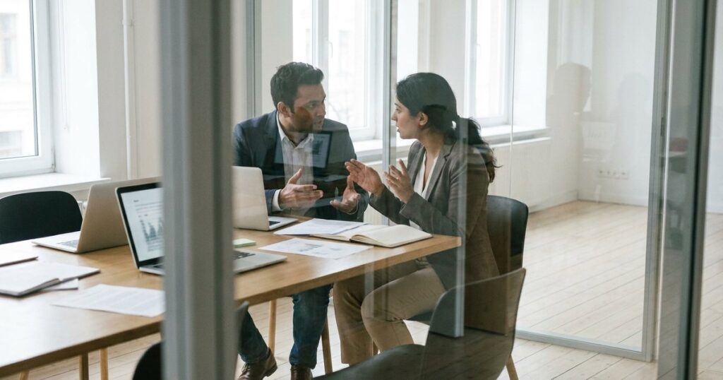Two colleagues in a startup office discussing financial strategies through a glass partition, related to balance sheets.